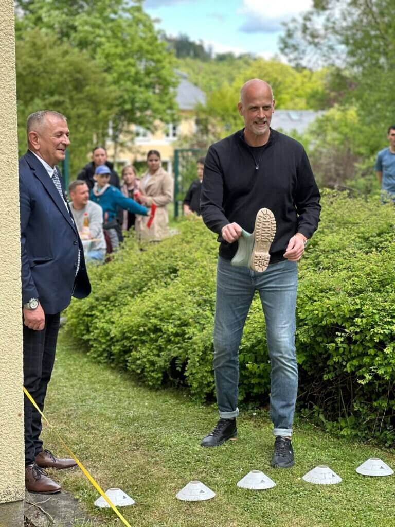 Ein Mann mit einem Gummistiefel in der Hand neben einem weiteren Mann in Anzug, Menschen im Hintergrund
