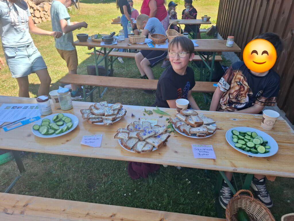 Kinder sitzen an Tischen, auf einem Tisch stehen 5 Teller mit Butterbroten oder Gurken