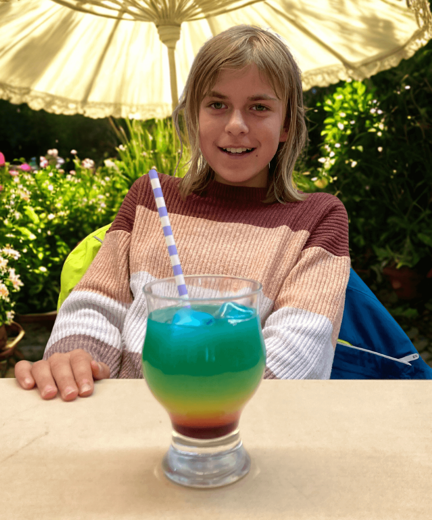 Ein Mädchen sitzt an einem Tisch, Hintergrund Garten und Sonnenschirm, auf dem Tisch steht ein bunter Cocktail mit Strohhalm