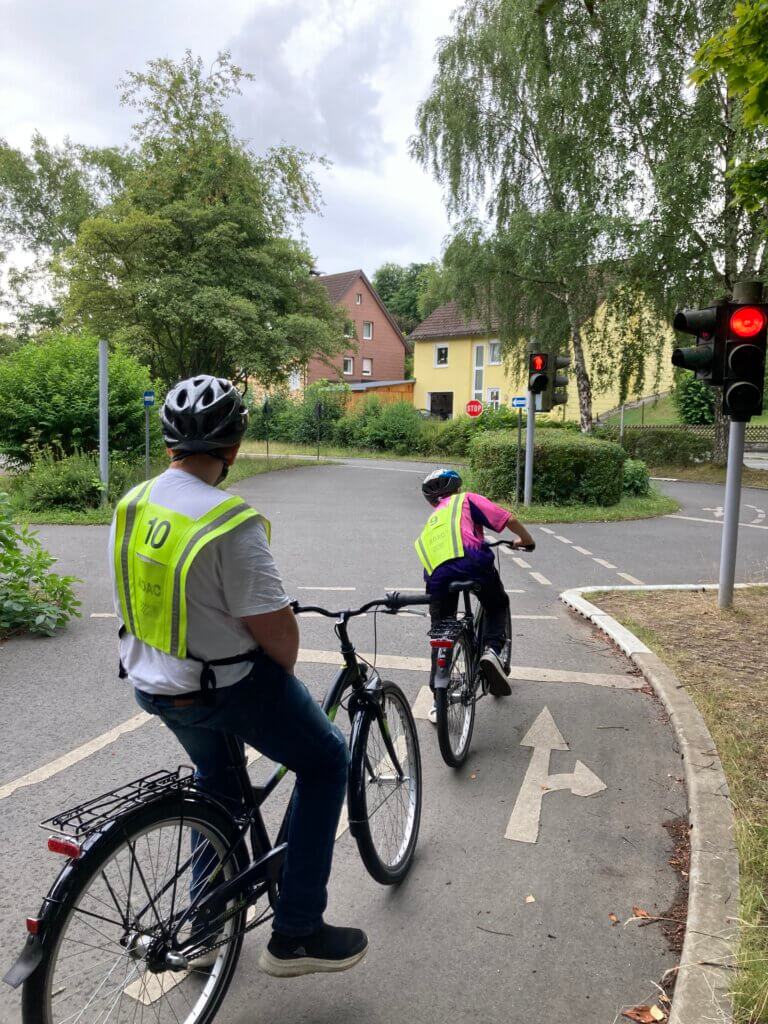 2 Kinder stehen am Verkehrsübungsplatz auf ihren Fahrrädern vor einer roten Ampel und warten.